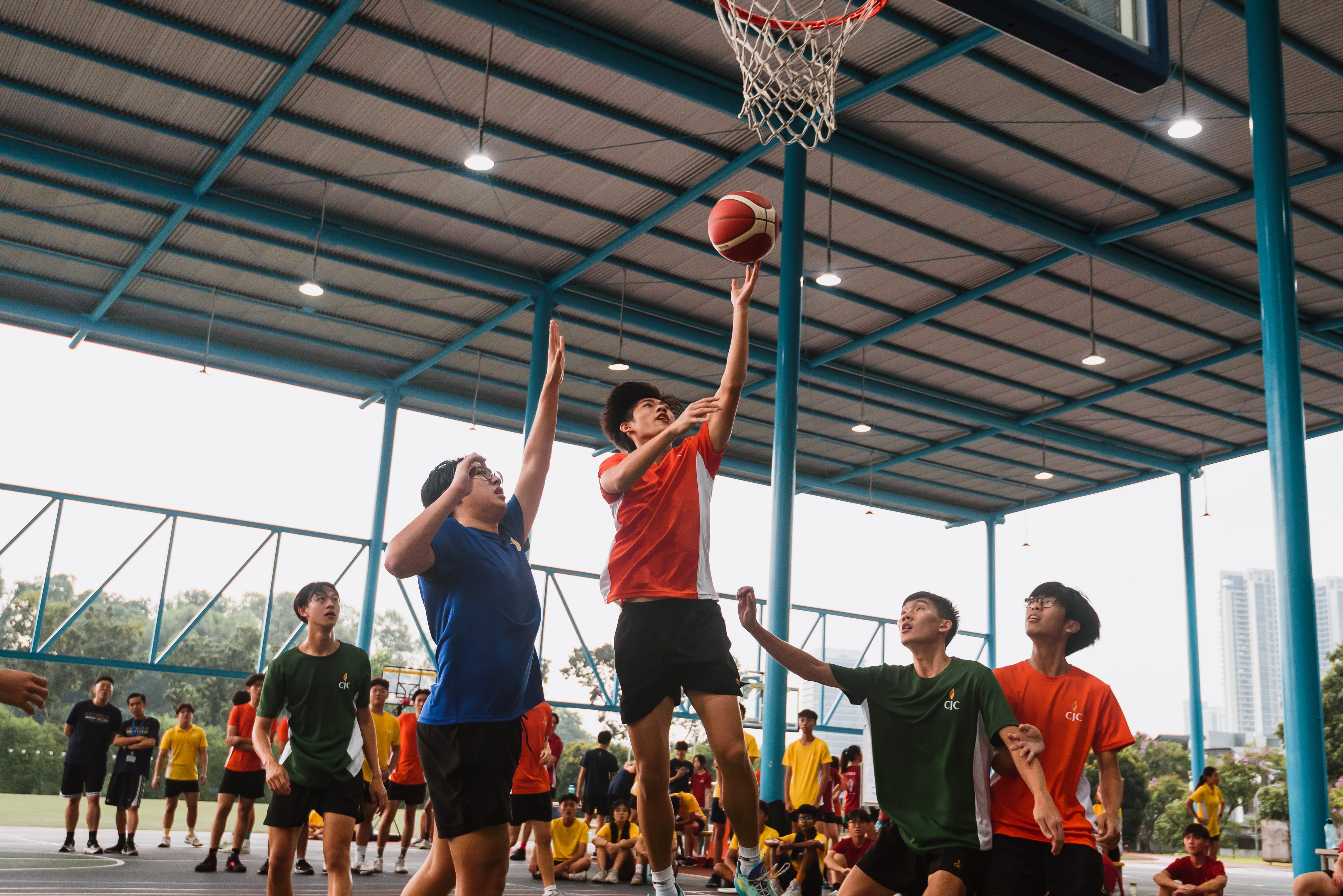 Indoor court with teens playing basketball; one jumps for a layup as others reach under the hoop.